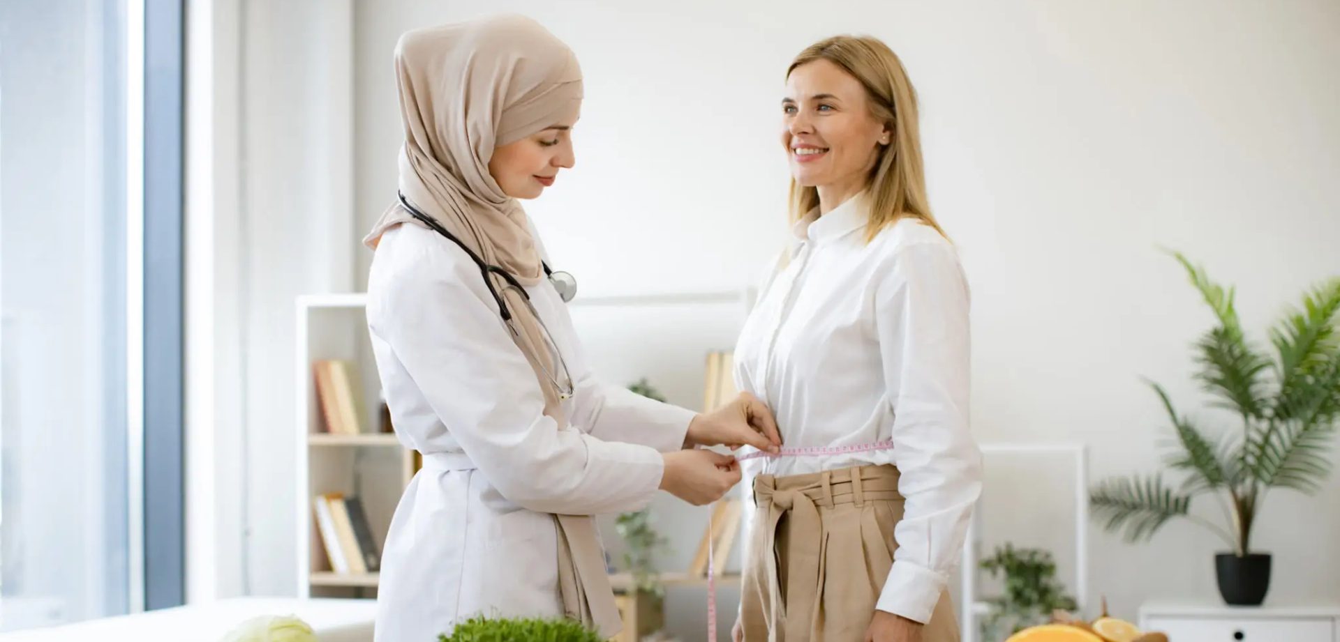 Two women, one in a hijab, adjusting the other's outfit in a bright room.