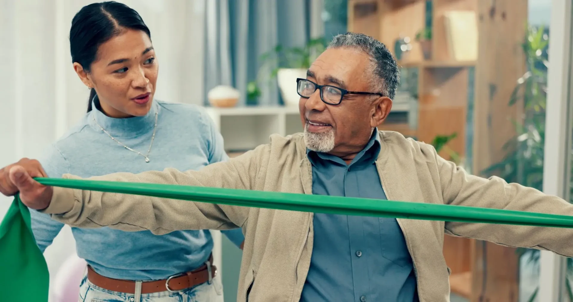 A young man helps an elderly man exercise with a resistance band.