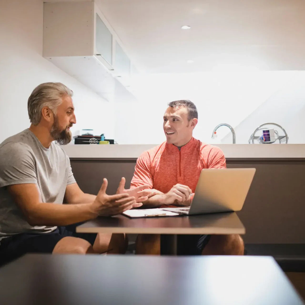 Two men discussing something while sitting at a kitchen table with a laptop.